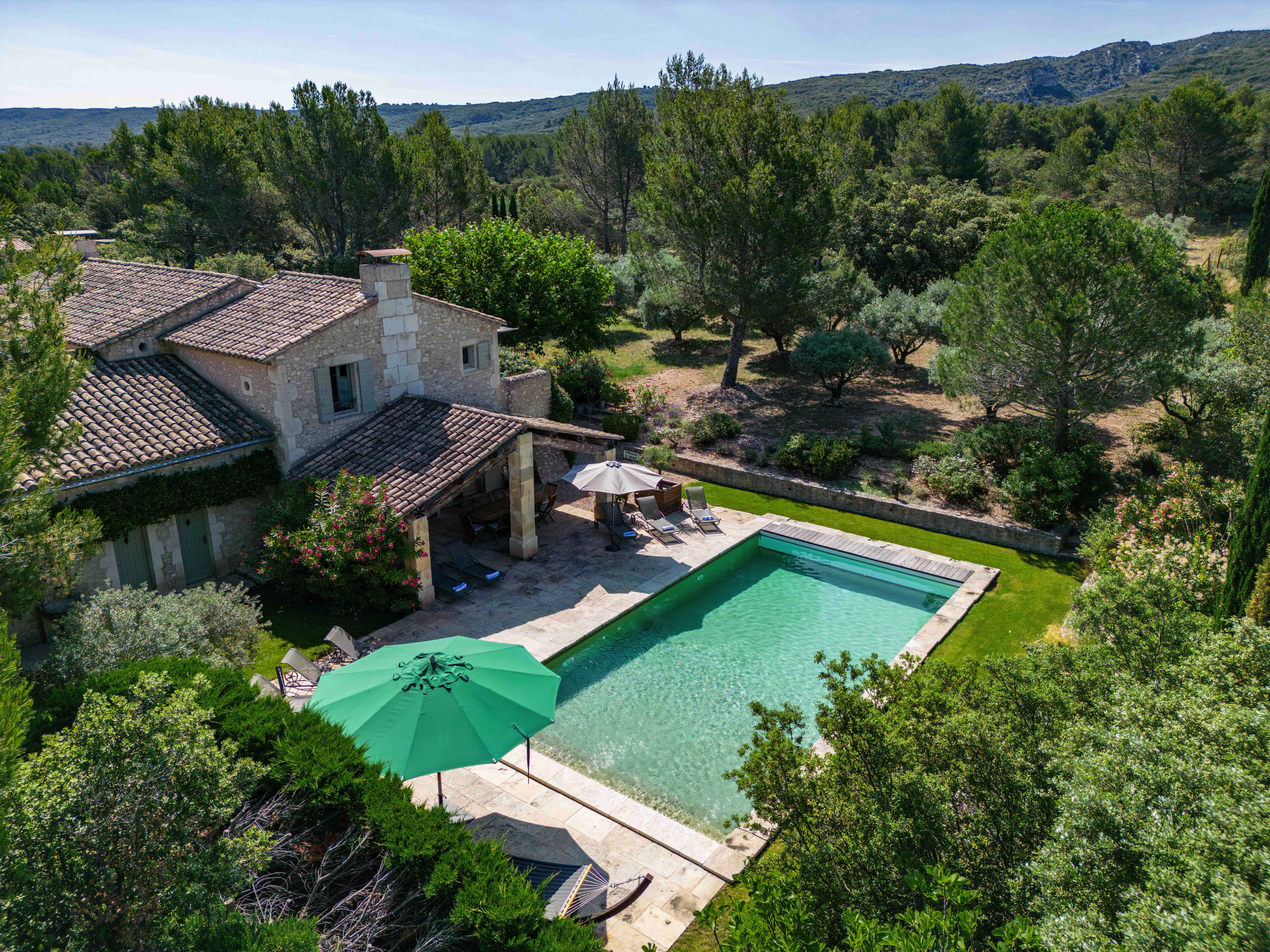 Aerial view of a countryside villa with a pool, surrounded by lush greenery and tall trees under a clear blue sky. A green umbrella and loungers are by the poolside.